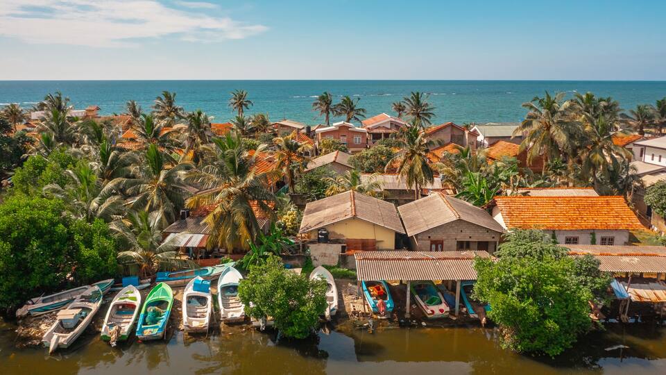 A coastal village with boats docked along a waterway, surrounded by palm trees and houses with red-tiled roofs.