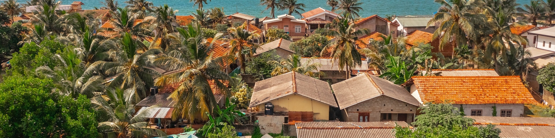 Uma vila costeira com barcos ancorados ao longo de um canal, cercada por palmeiras e casas com telhados vermelhos.