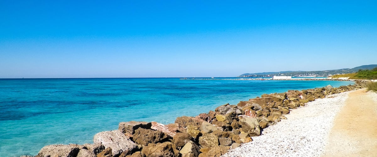A coastal landscape with clear turquoise waters, a rocky shoreline, and distant buildings on the horizon.