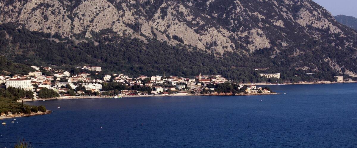 A coastal town with buildings along the shore, a large mountain in the background, and a clear blue sea.