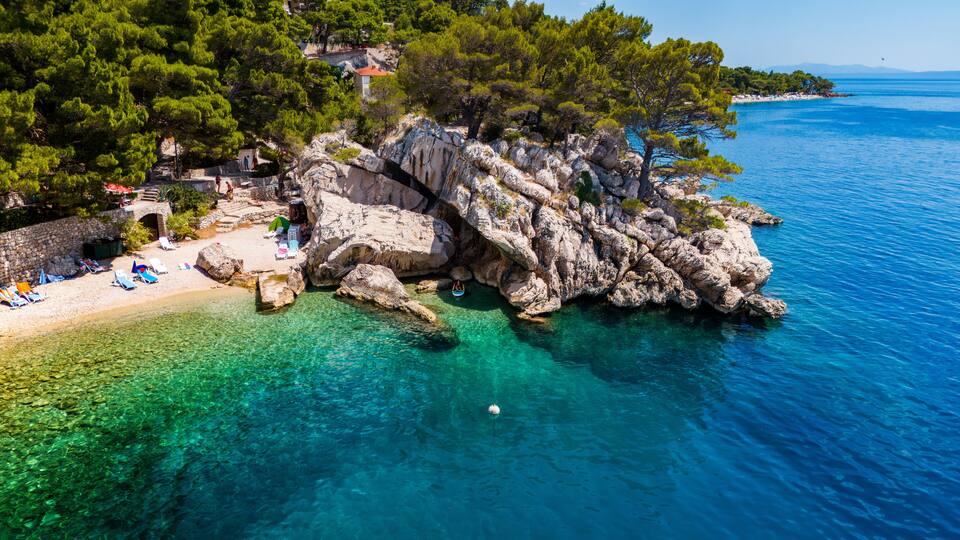 A rocky coastline with clear turquoise waters, a sandy beach with umbrellas, and lush green trees.