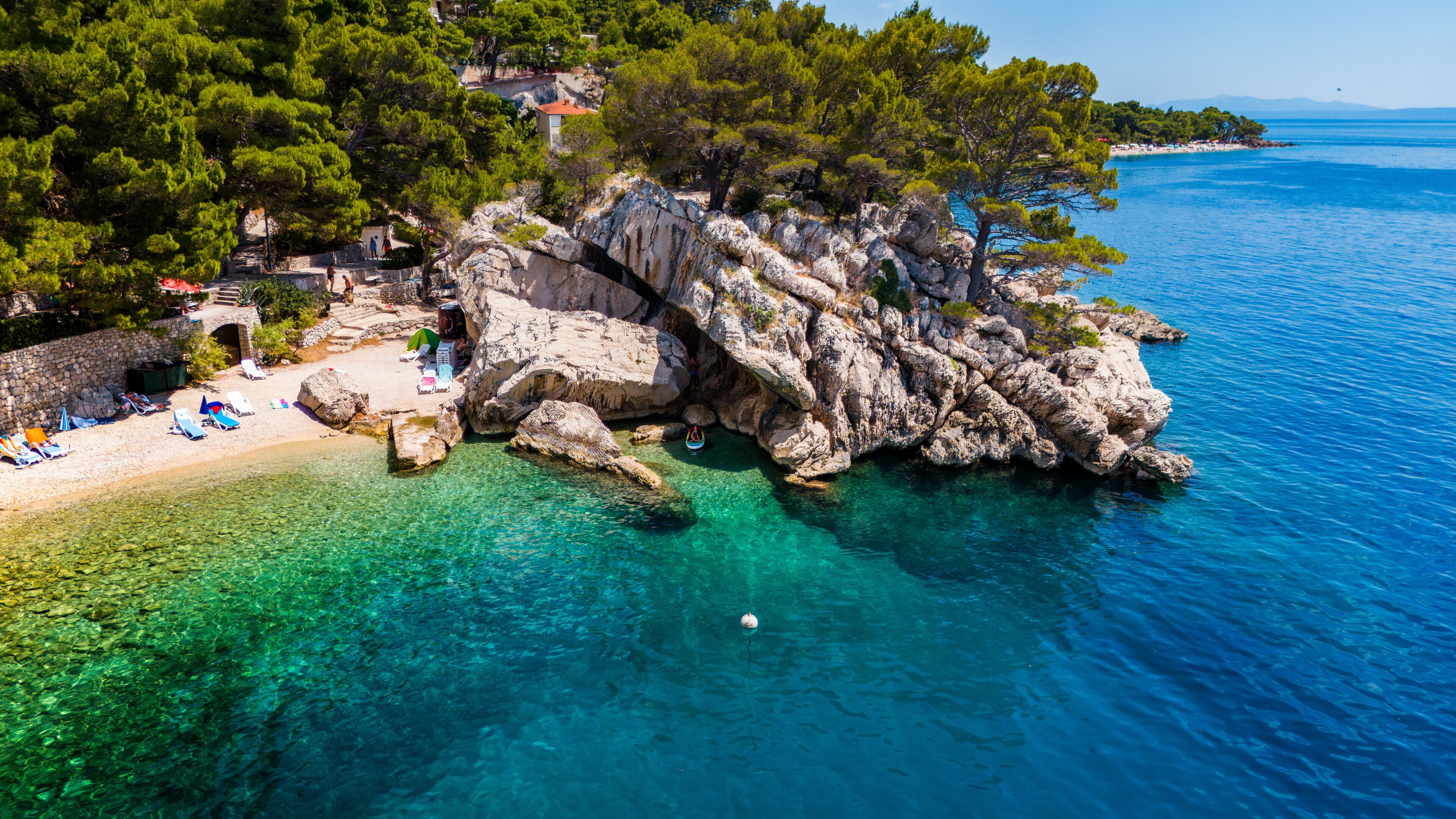 A rocky coastline with clear turquoise waters, a sandy beach with umbrellas, and lush green trees.