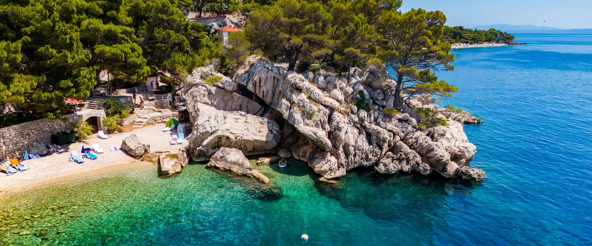 A rocky coastline with clear turquoise waters, a sandy beach with umbrellas, and lush green trees.