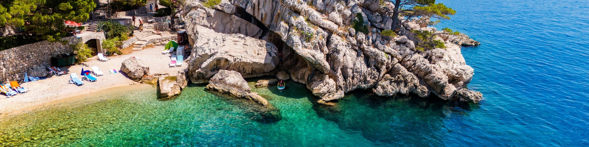 A rocky coastline with clear turquoise waters, a sandy beach with umbrellas, and lush green trees.