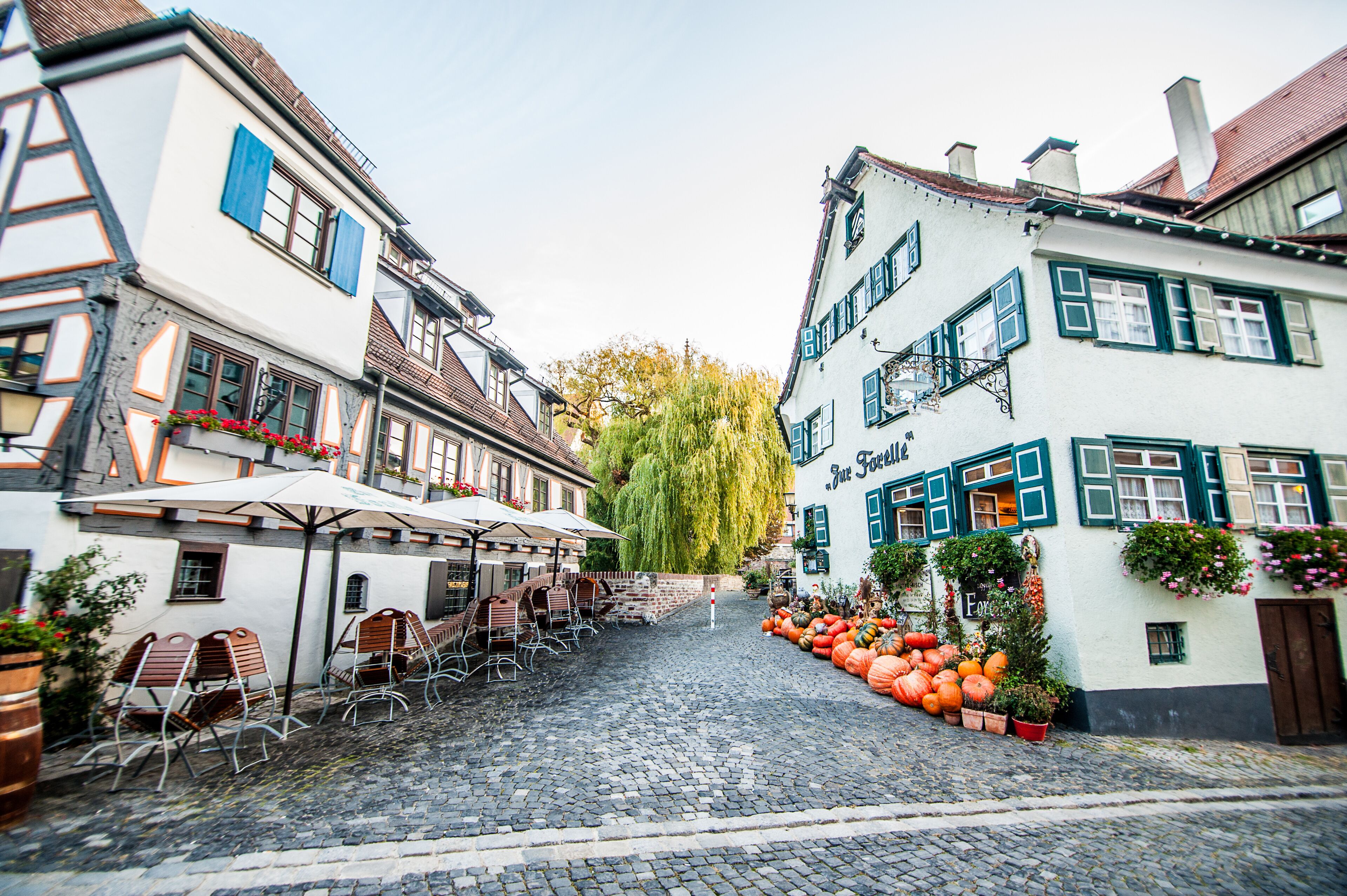 A cobblestone street lined with traditional European buildings, outdoor seating, and autumnal decorations.