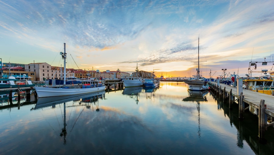 A marina with boats docked at wooden piers, buildings in the background, and a sunset over the water.