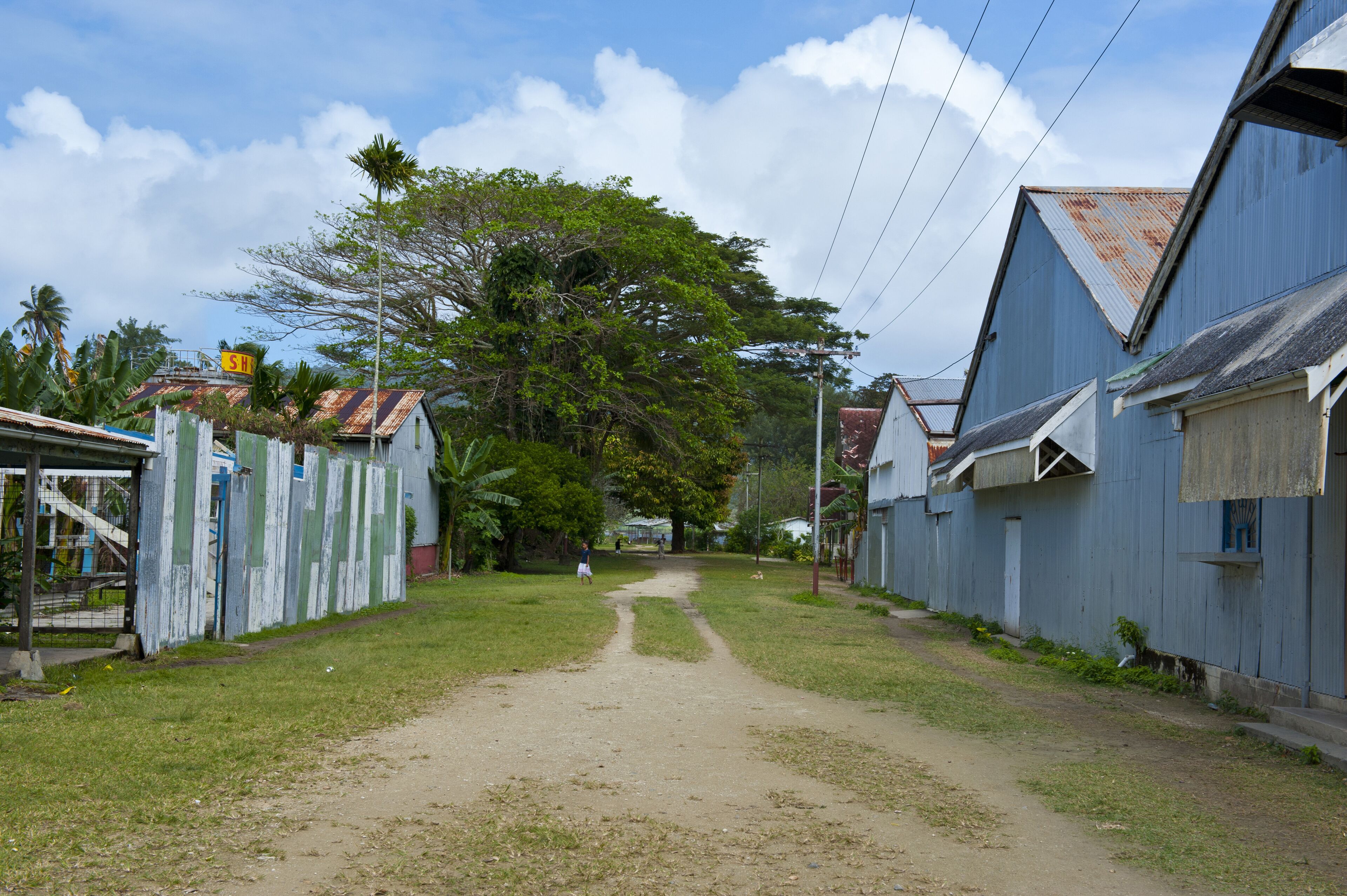 A narrow dirt path flanked by buildings with corrugated metal roofs, surrounded by greenery and a clear sky.