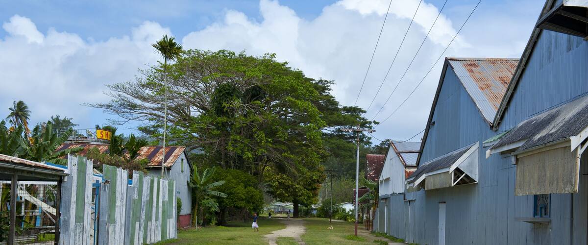 A narrow dirt path flanked by buildings with corrugated metal roofs, surrounded by greenery and a clear sky.