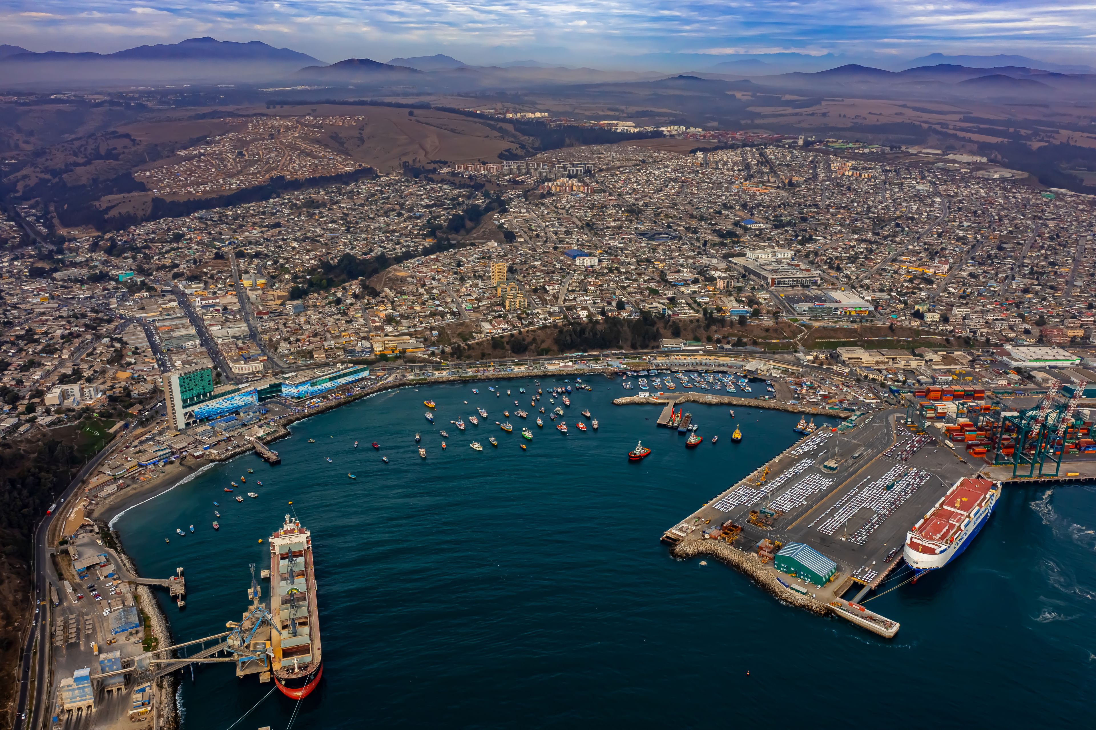 A coastal cityscape with a harbor, ships, and a mountainous backdrop.
