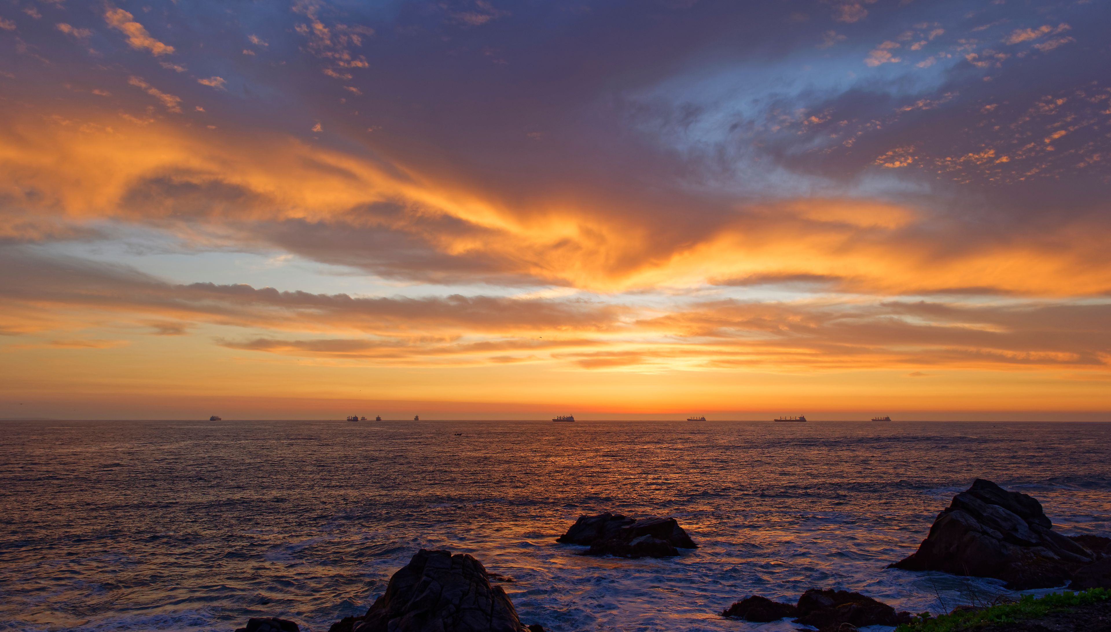 A sunset over the ocean with ships and rocky outcrops.