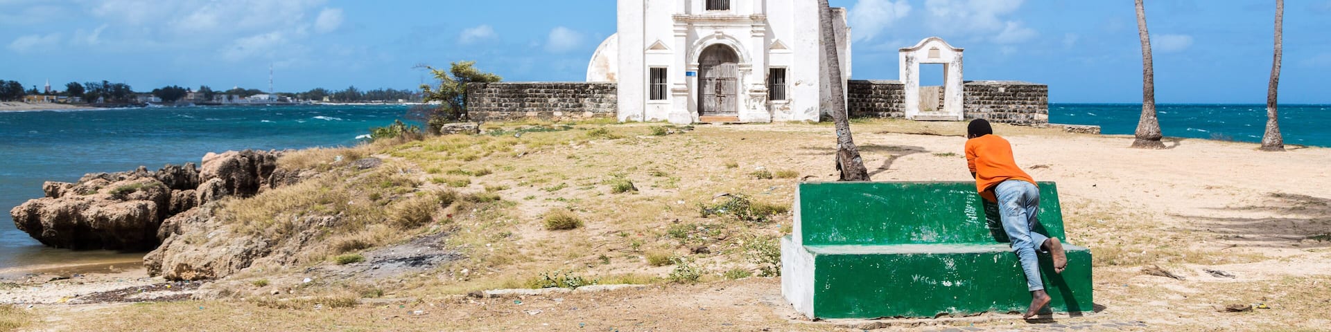 A man in orange t-shirt looking at Church fortress of San Antonio on Mozambique island, with three palm trees on sand. Indian ocean coast, Mozambique.