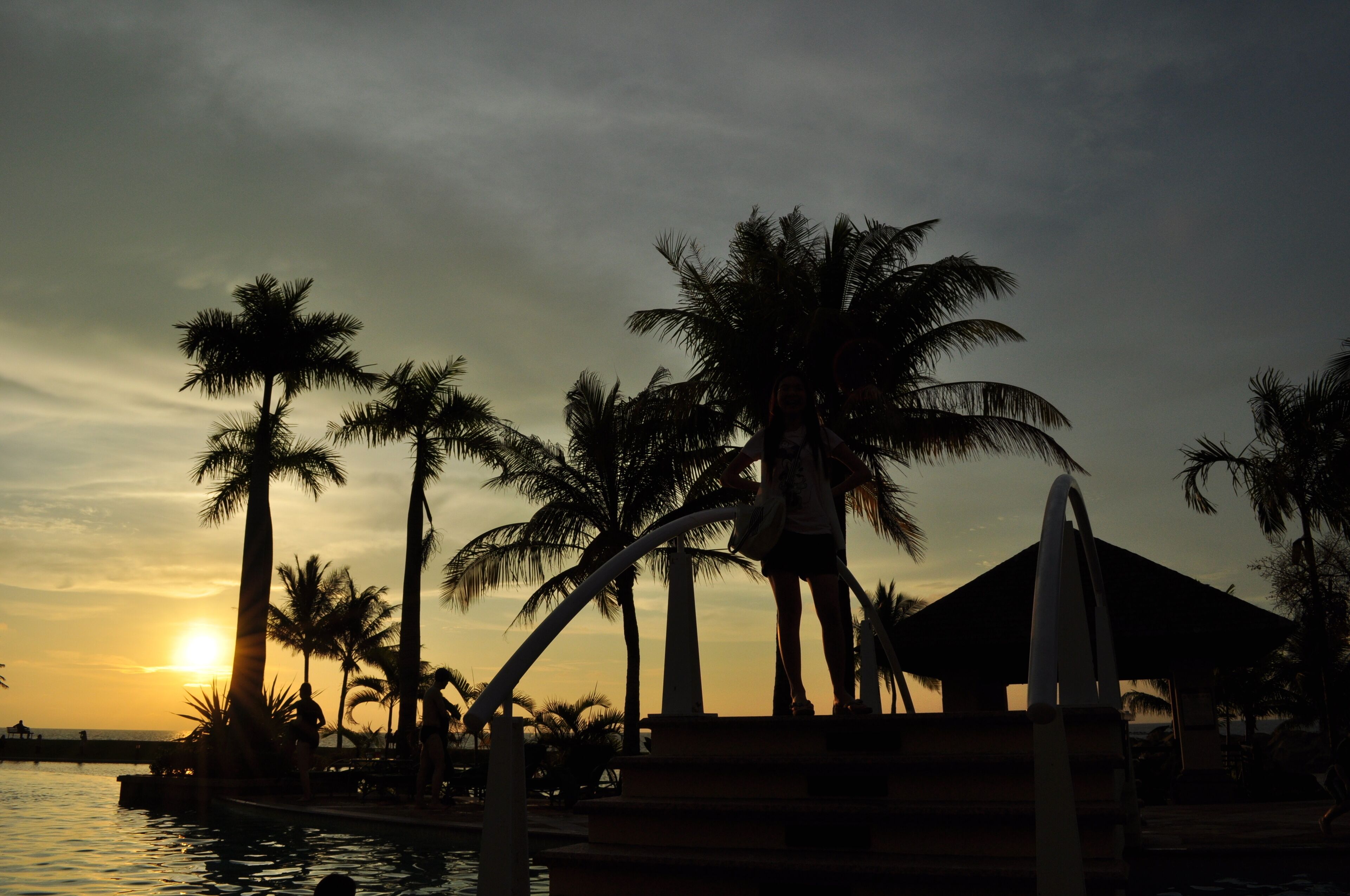 A sunset over a beach with palm trees and a pool.
