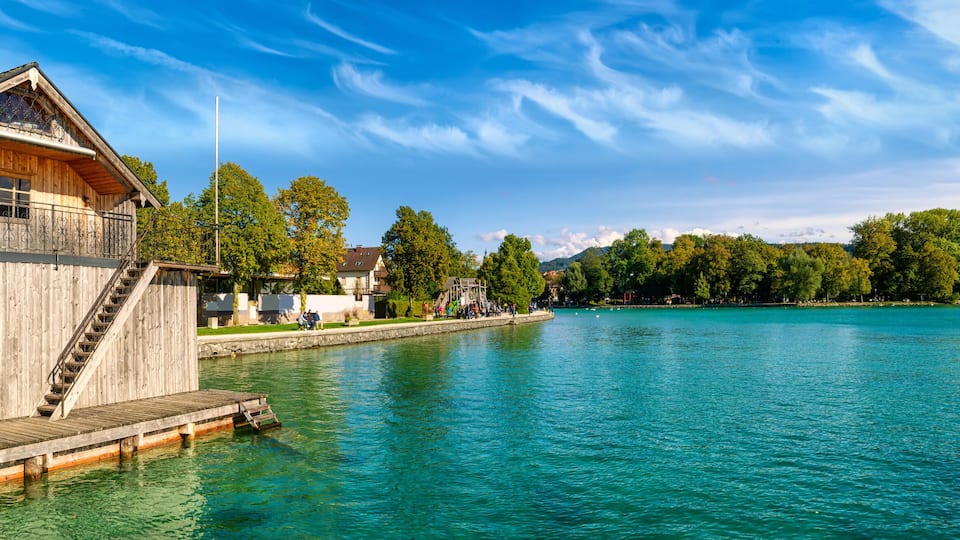 A wooden house with a balcony on stilts by a calm lake, surrounded by trees and mountains.