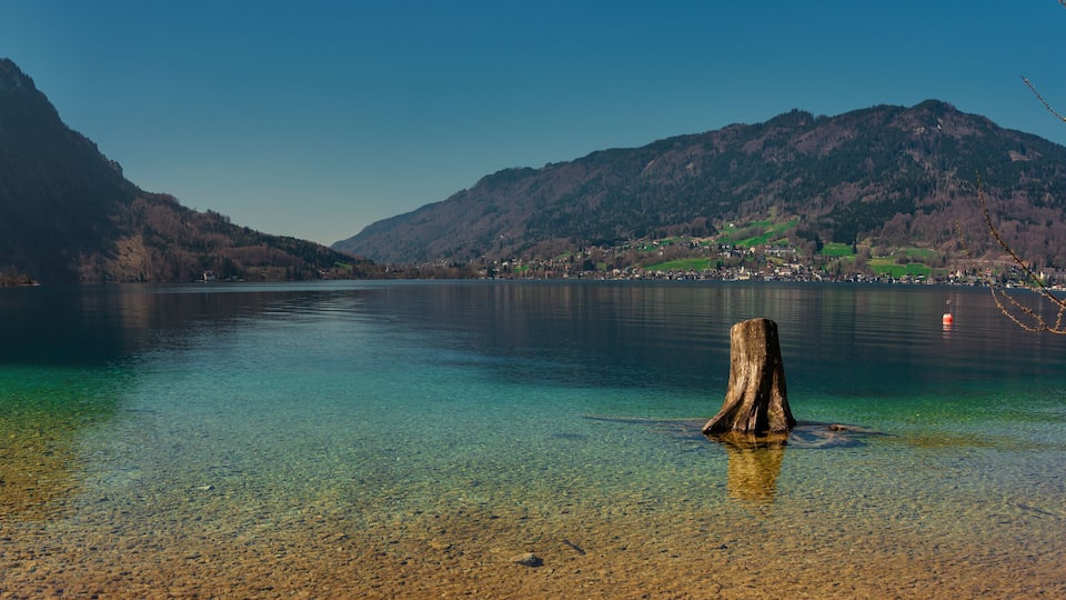 A clear lake with a tree stump in the foreground, surrounded by mountains and a small town in the background.