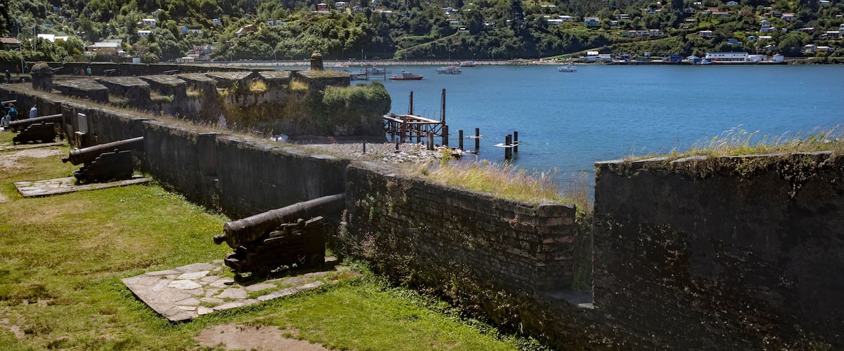 A coastal fort with cannons, a calm harbor with boats, and a hillside village in the background.