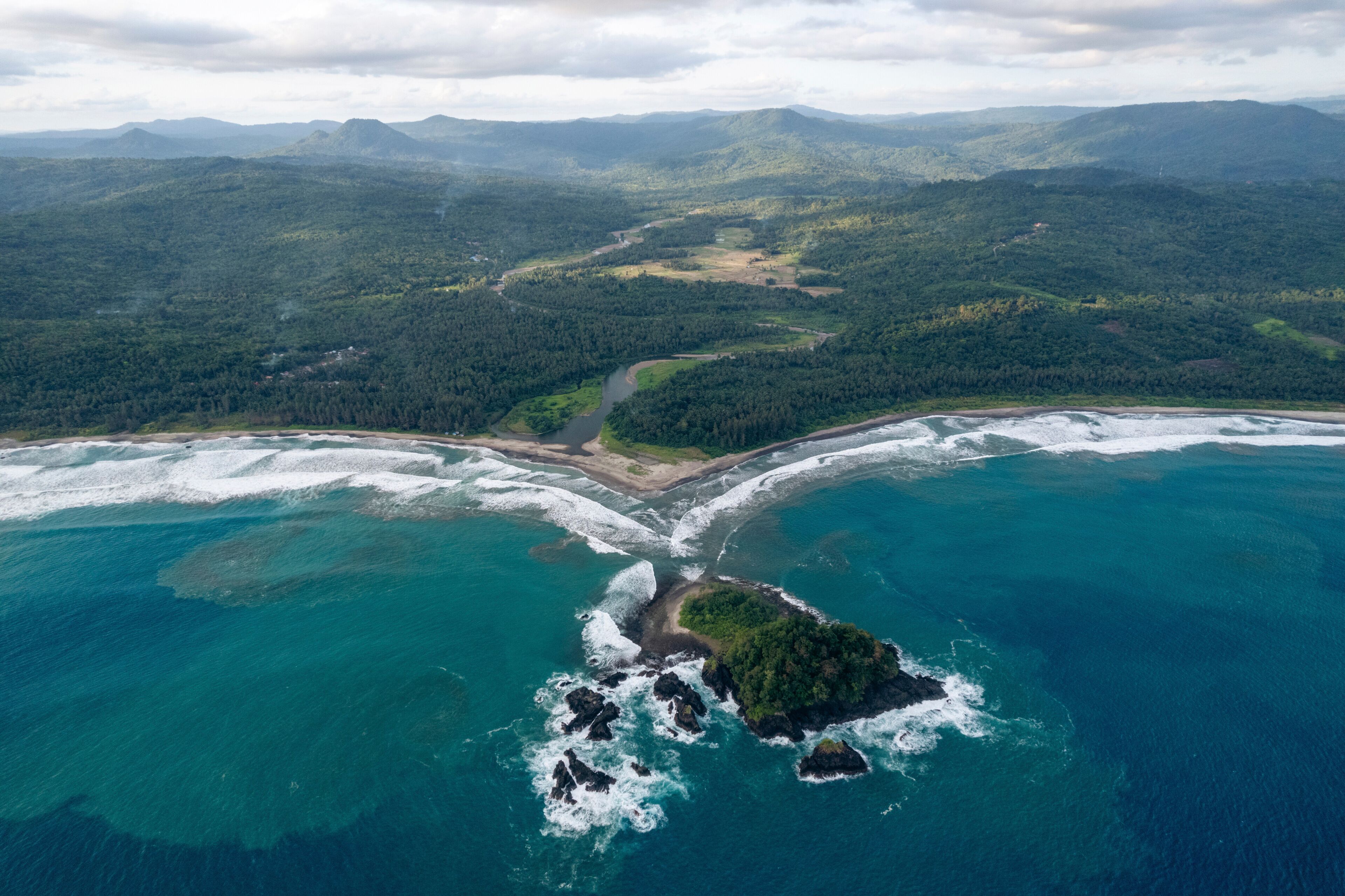 Aerial view of a coastline with waves, lush green hills, and a small island. South Nias, North Sumatra, Indonesia