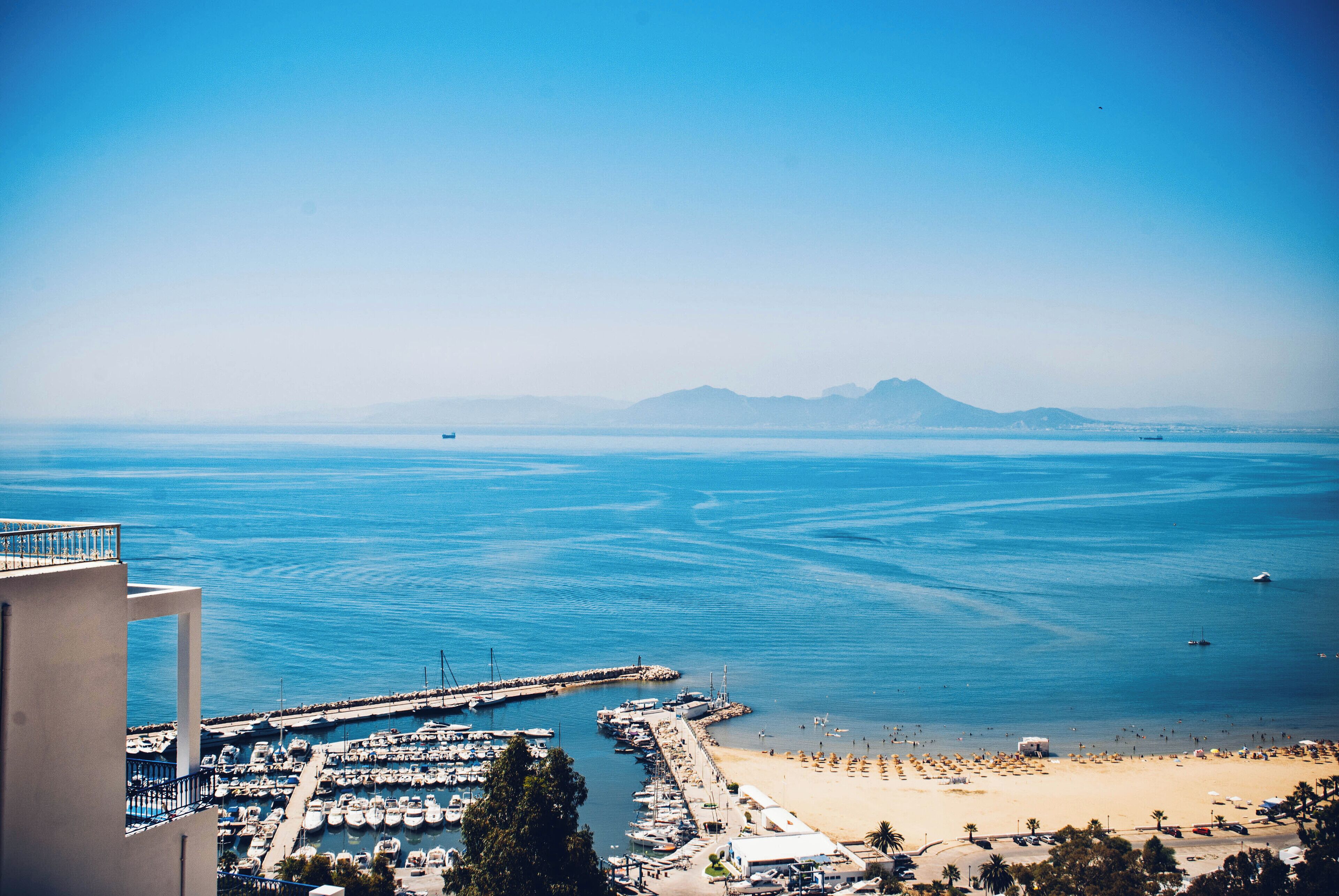 A coastal marina with boats, a beach, and mountains in the distance.