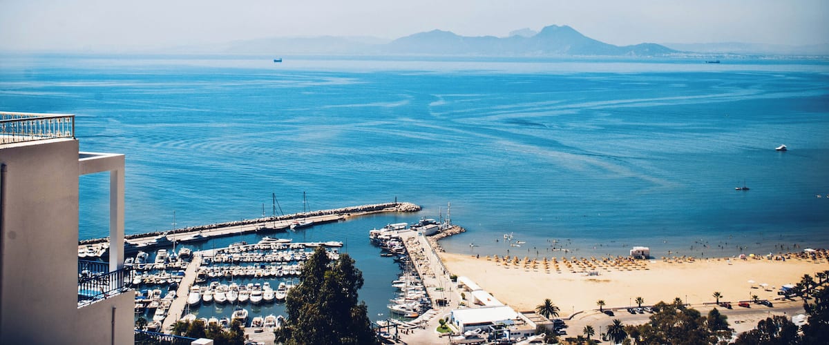 A coastal marina with boats, a beach, and mountains in the distance.