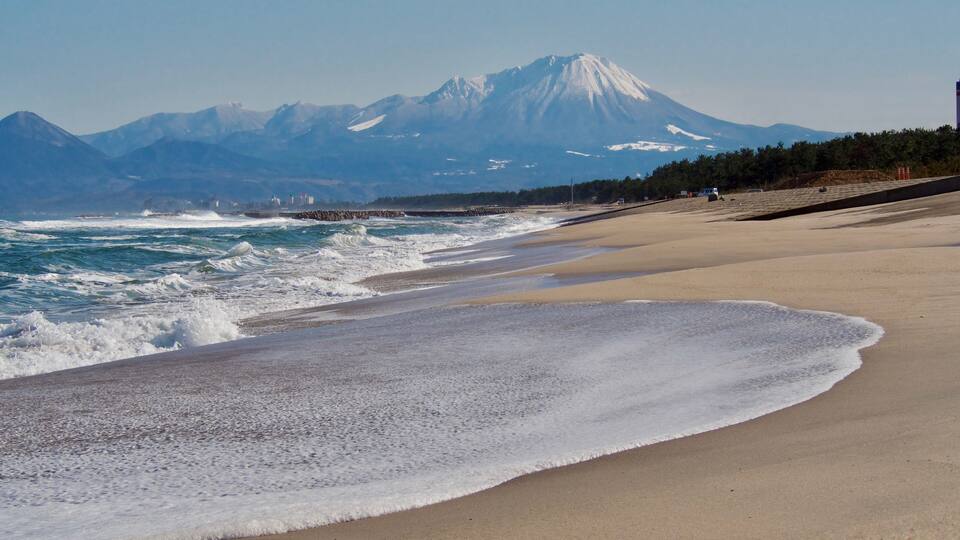 A beach with waves, a distant snow-capped mountain, and a rocky shoreline.