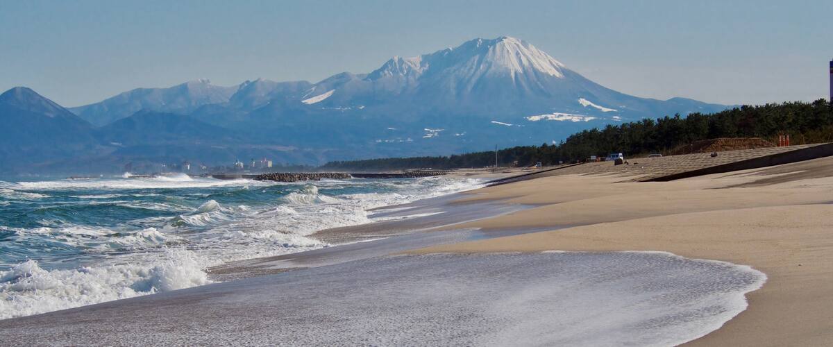 A beach with waves, a distant snow-capped mountain, and a rocky shoreline.