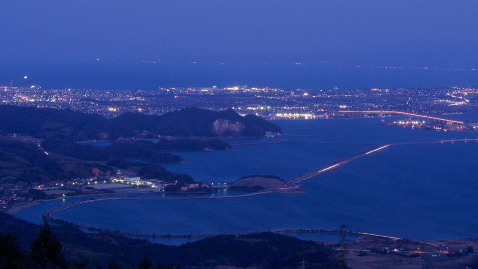 A coastal cityscape at dusk with a prominent bridge and a large body of water.