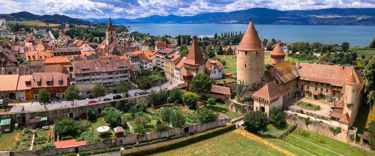 A historic town with medieval architecture, including towers and a church, surrounded by a lake and mountains.