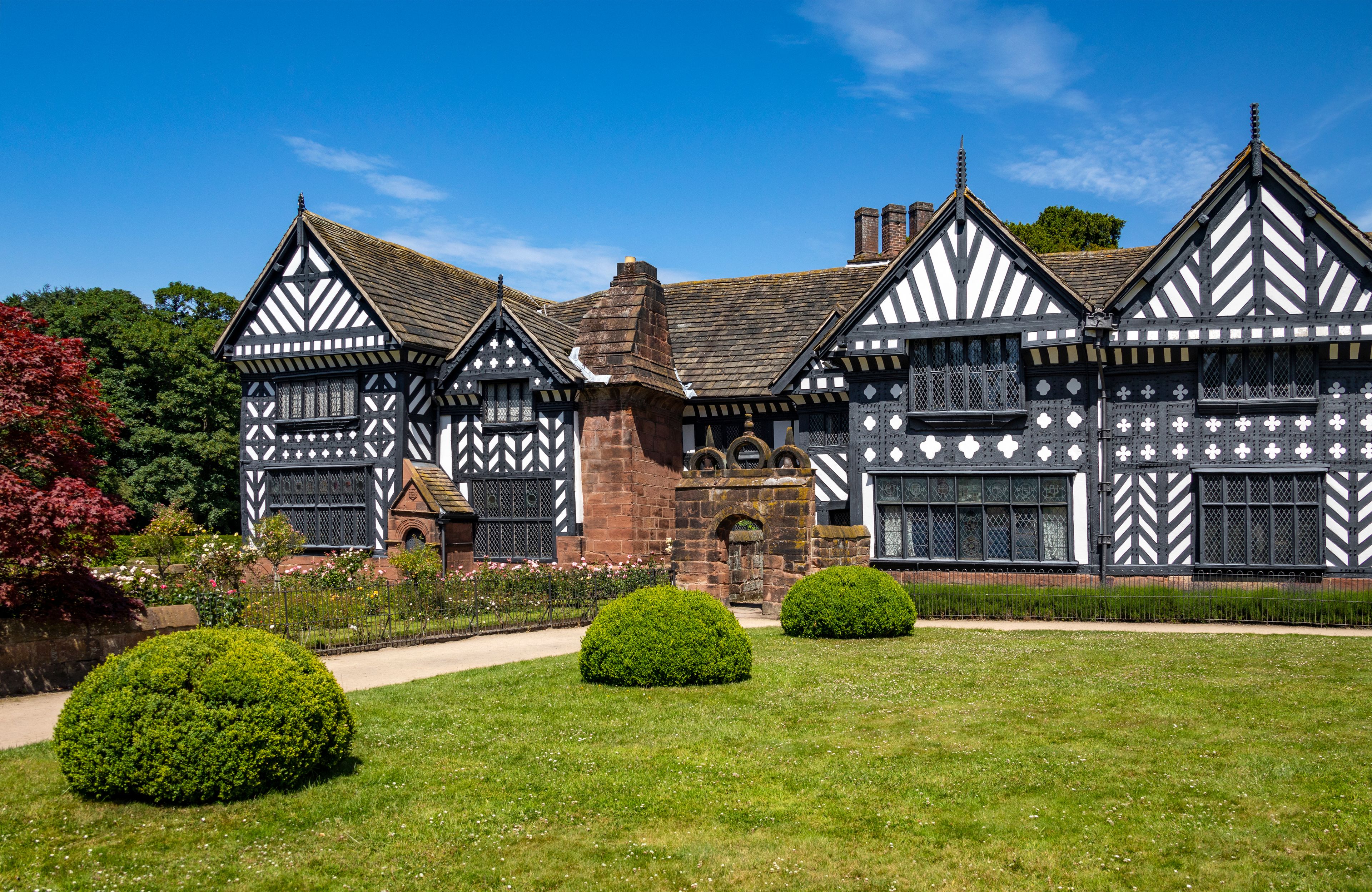 Un edificio de estilo Tudor con entramado de madera en blanco y negro, rodeado de jardines cuidados y un cielo azul despejado.