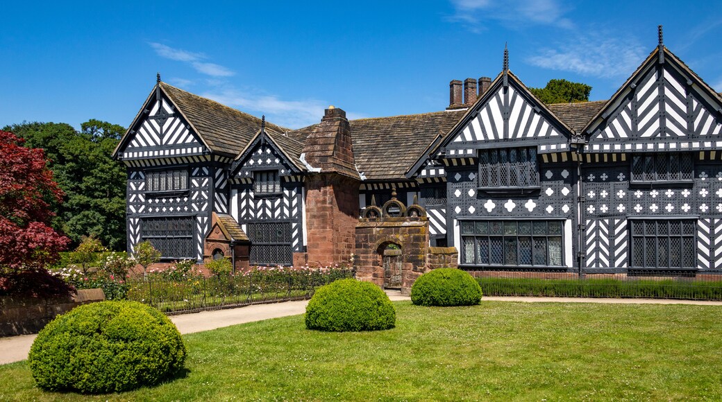 Un edificio de estilo Tudor con entramado de madera en blanco y negro, rodeado de jardines cuidados y un cielo azul despejado.