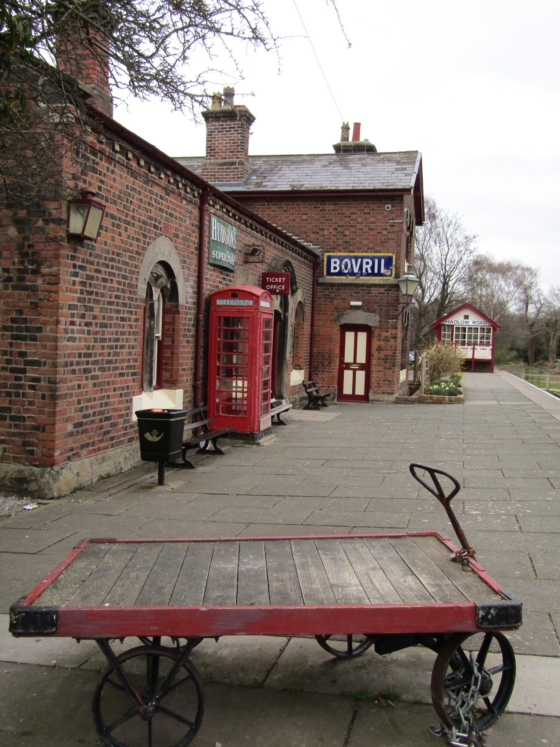 A red telephone booth, a black trash bin, and a wooden cart in front of a brick building with a sign that reads "BOVRIL".