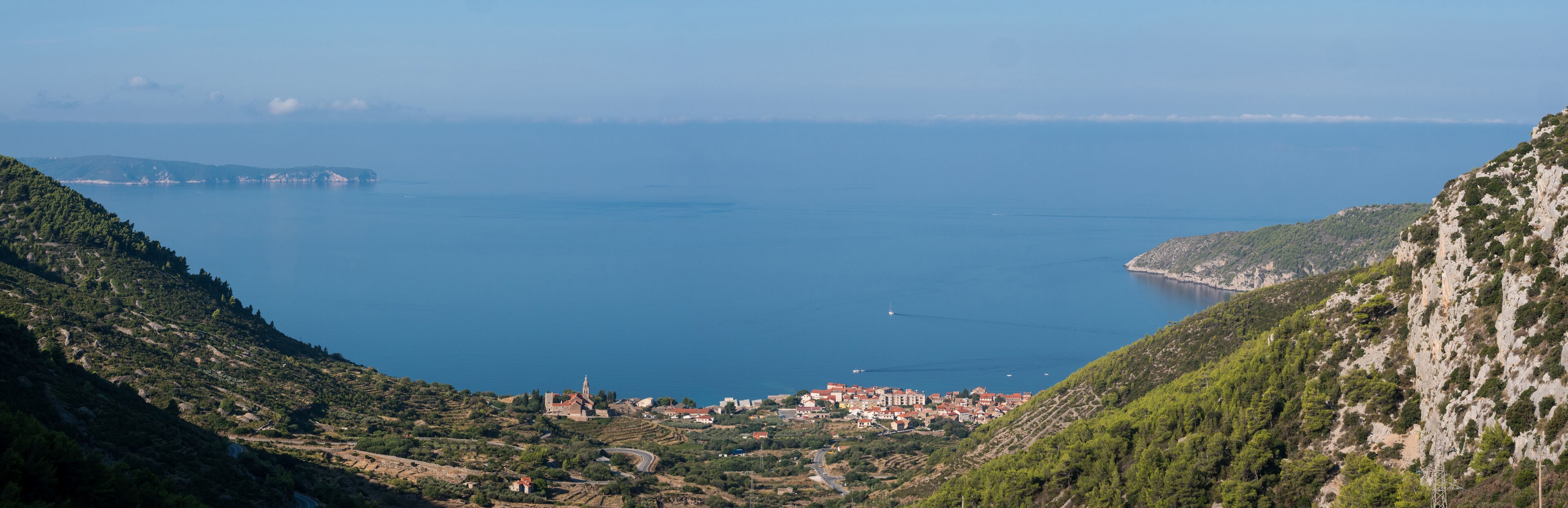 A coastal town nestled between hills, with a clear view of the sea and a distant island.