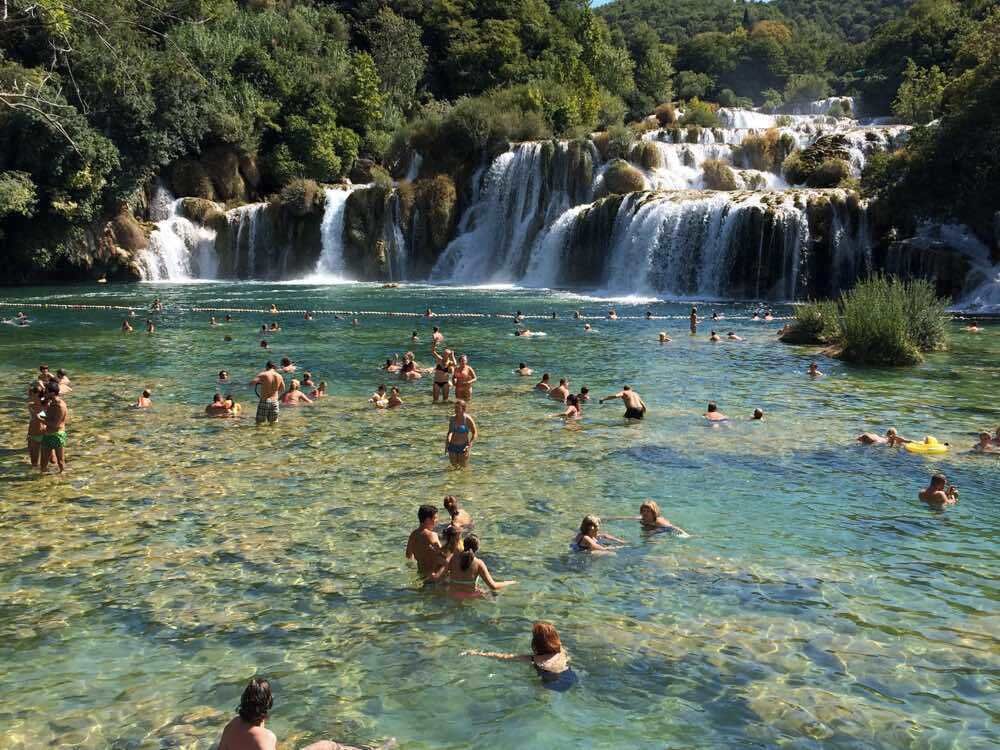 A group of people swimming in a clear river with a waterfall in the background.