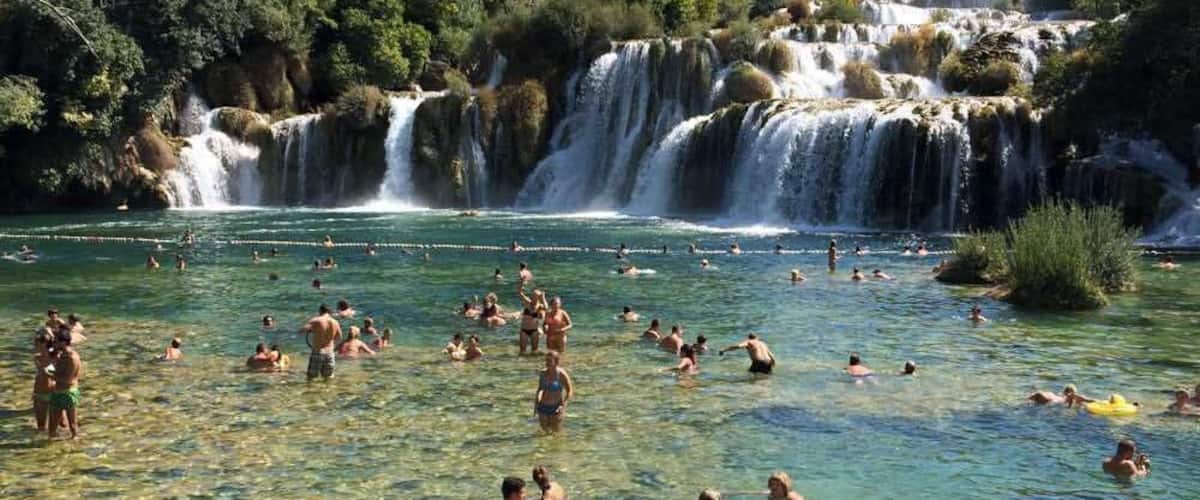 A group of people swimming in a clear river with a waterfall in the background.
