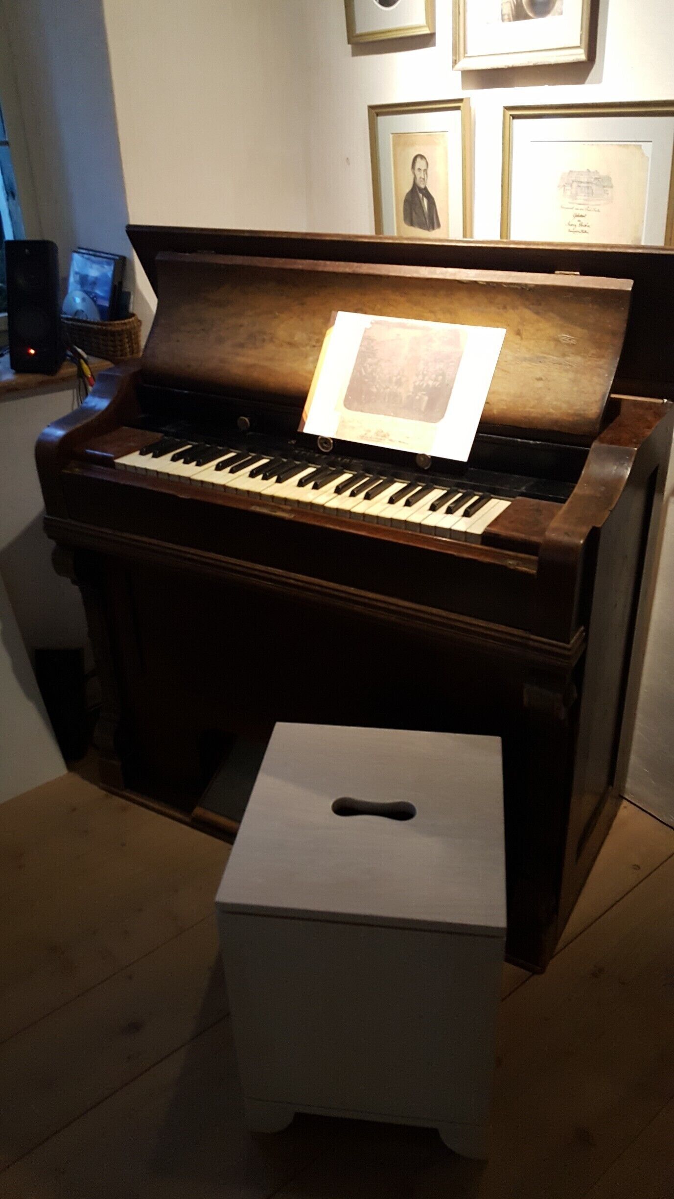 A grand piano with sheet music on it, a small stool in front, and framed pictures on the wall.