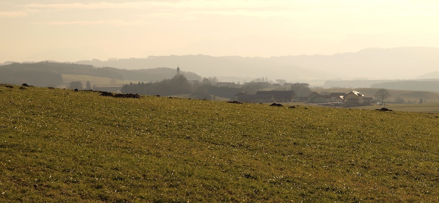 Un paysage rural brumeux, avec des collines ondoyantes, des maisons éparpillées et un clocher d’église imposant.