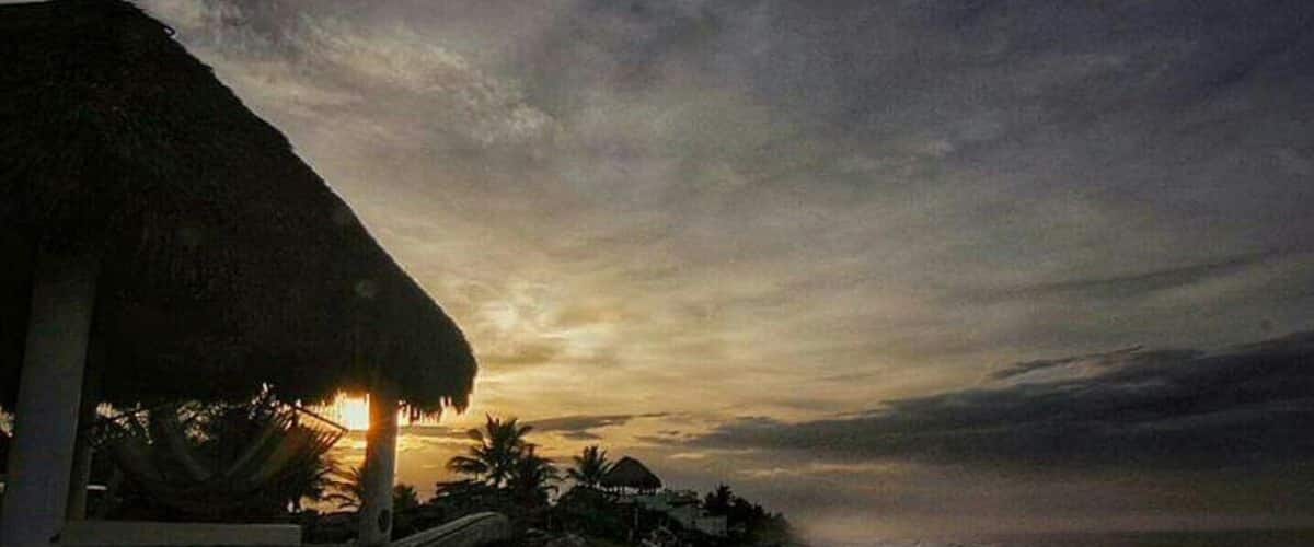 A beach at sunset with a thatched-roof hut and palm trees.