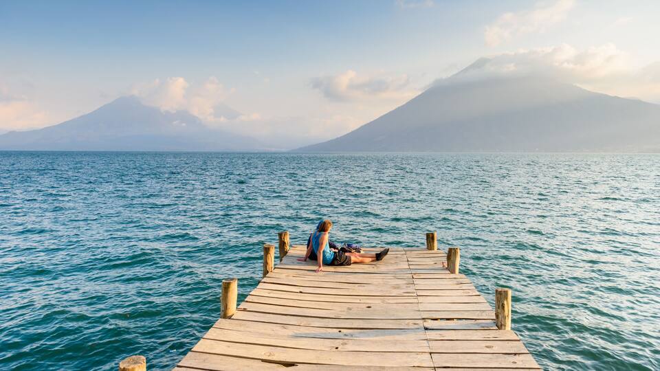 A wooden pier extending into a calm sea with a person sitting at the end, overlooking a mountain range.