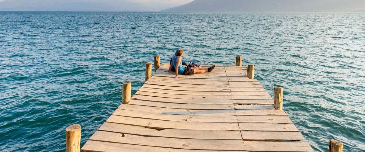 A wooden pier extending into a calm sea with a person sitting at the end, overlooking a mountain range.