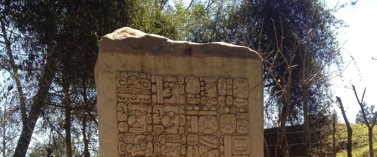 A stone monument with carved symbols and text, surrounded by trees and a fence.