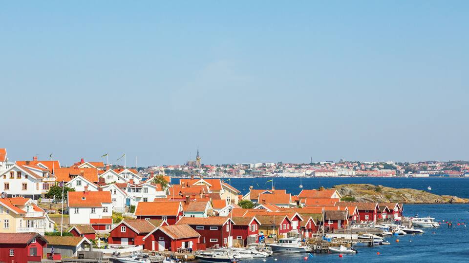 A coastal town with red and white houses, a harbor with boats, and a distant city skyline.