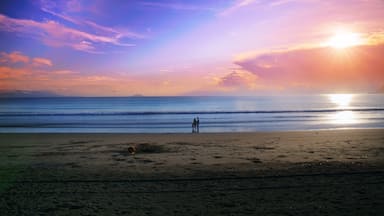 A beach with a sunset, ocean waves, and two people standing on the sand.