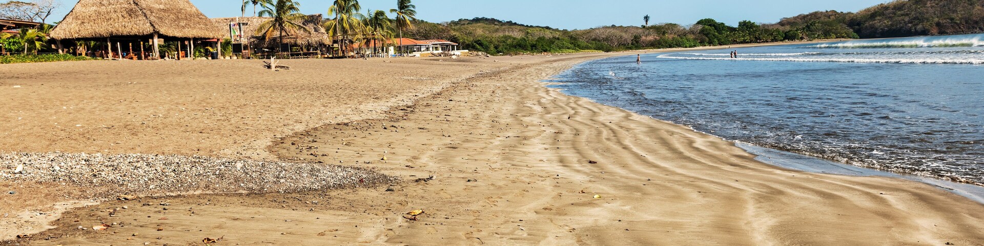 A sandy beach with footprints, palm trees, and thatched-roof huts.