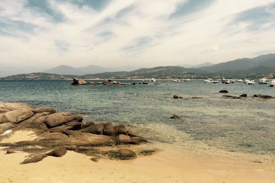 A beach with clear water, rocks, and boats in the distance.