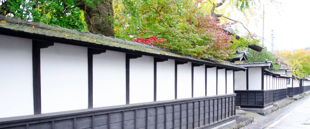 A traditional Japanese stone wall with white panels and black trim, lined with trees and autumn leaves.