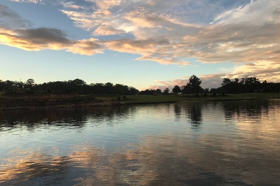 A calm lake with reflections of the sky and trees, surrounded by a grassy area and trees.
