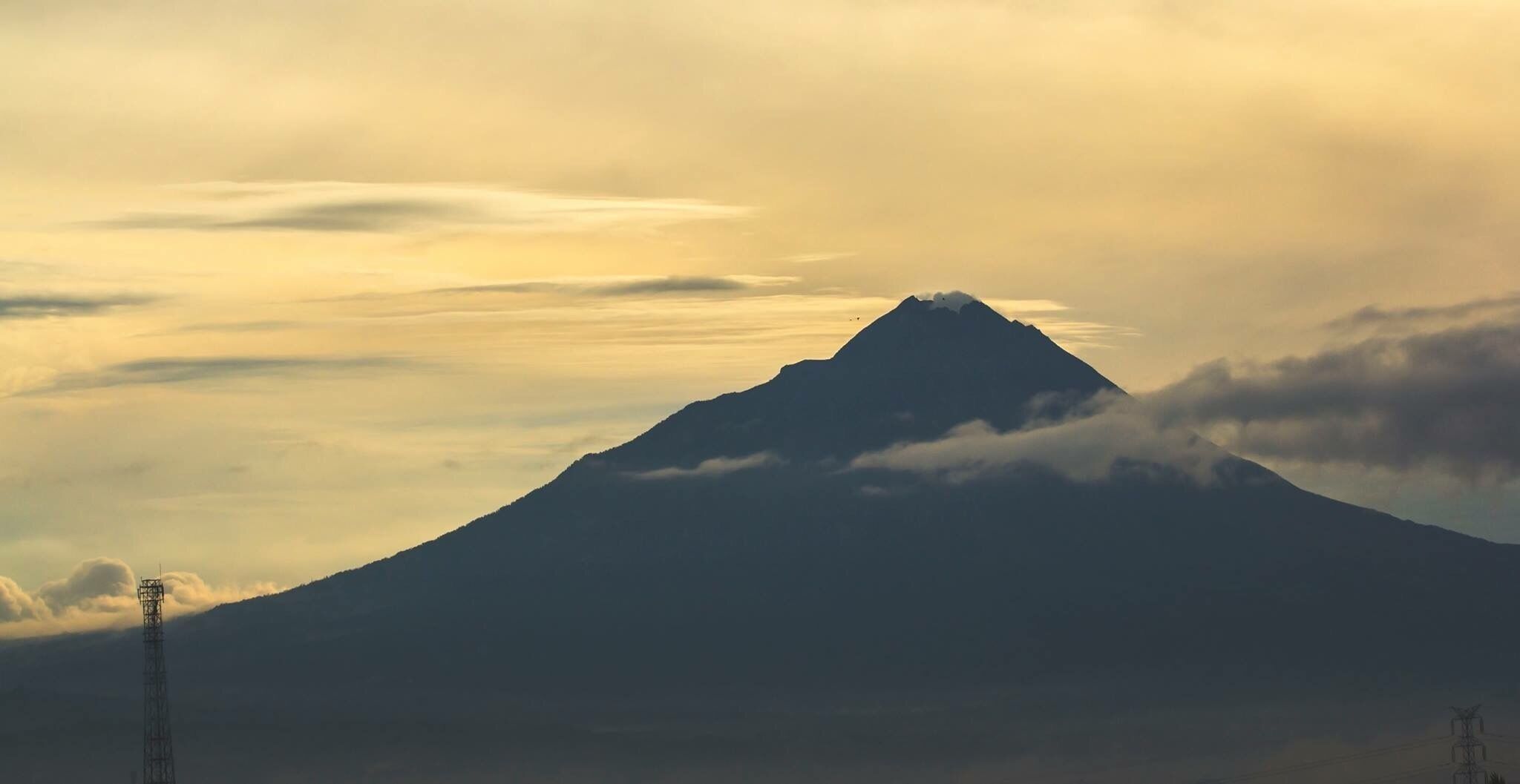 A mountain with a peak, surrounded by clouds and a power line tower.