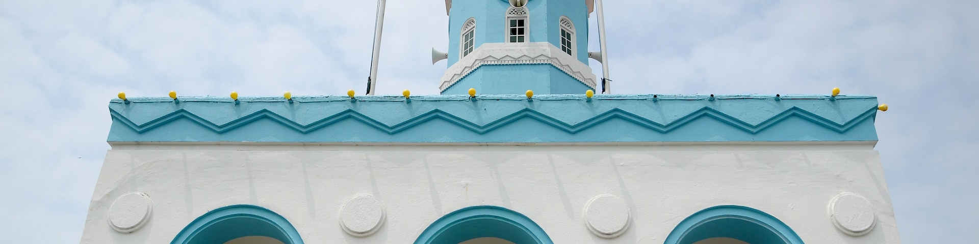 Un edificio con fachada azul y blanca, una torre de campanas y dos banderas ondeando en la parte superior.