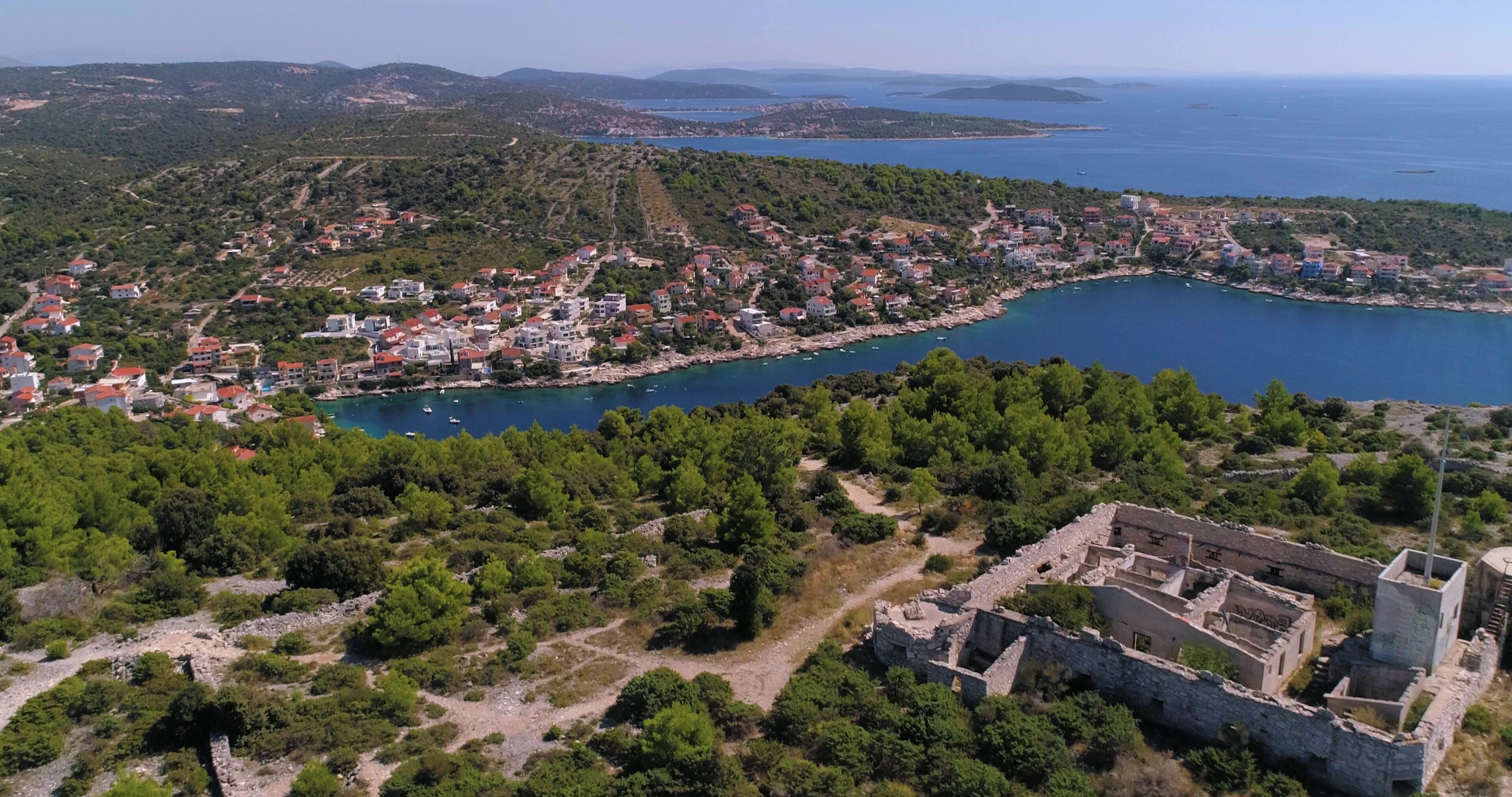 An aerial view of a coastal town with a historic fortress, dense greenery, and a clear blue sea.