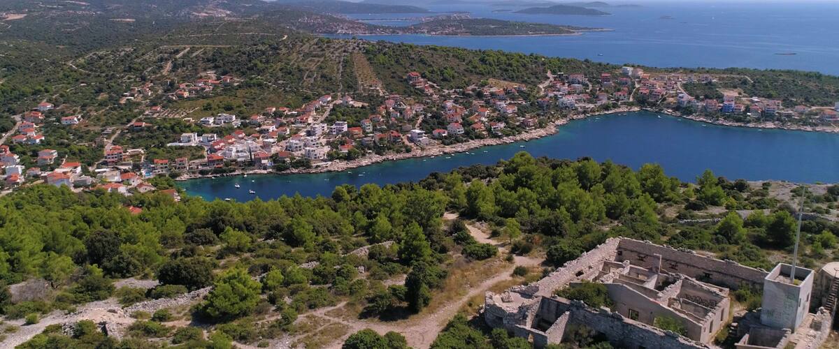 An aerial view of a coastal town with a historic fortress, dense greenery, and a clear blue sea.
