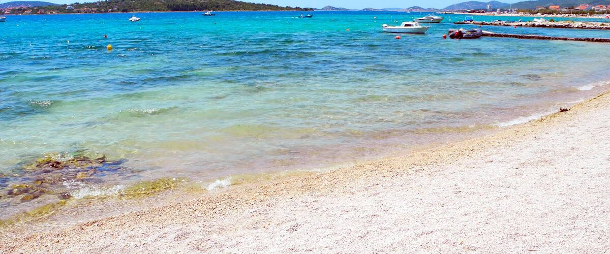 A beach with clear turquoise water, a sandy shore, and boats in the distance.