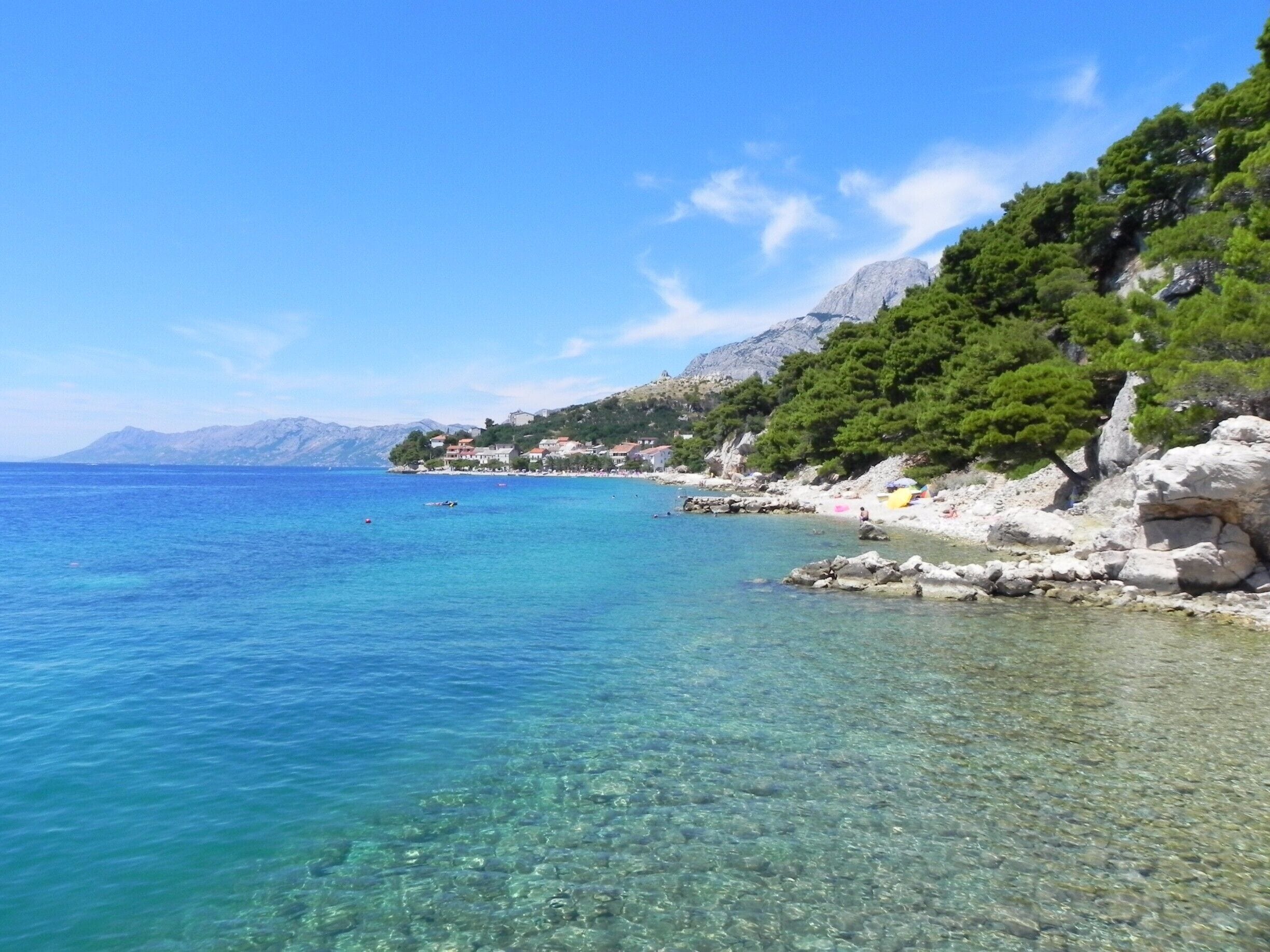 A clear coastal area with rocky shores, clear blue waters, and a backdrop of mountains and greenery.